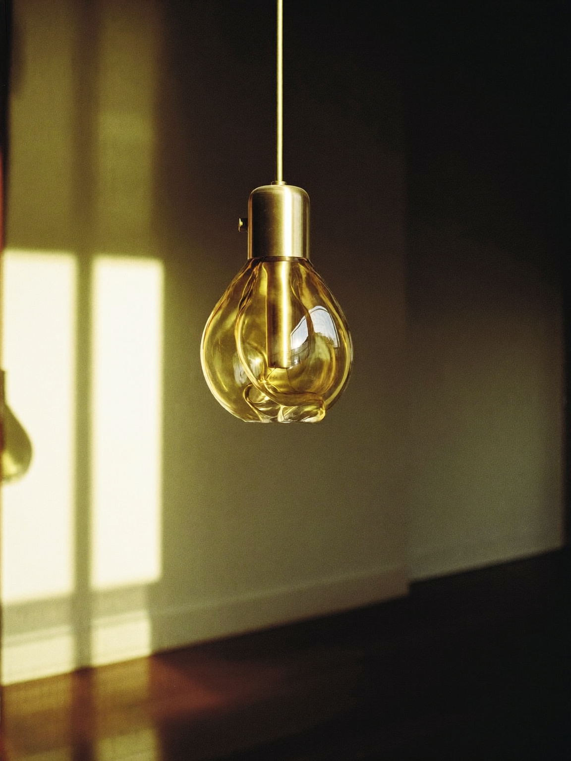 Lampada a sospensione scultorea in ottone e vetro soffiato ambrato, suspended in un interno milanese in penombra con pareti in gesso