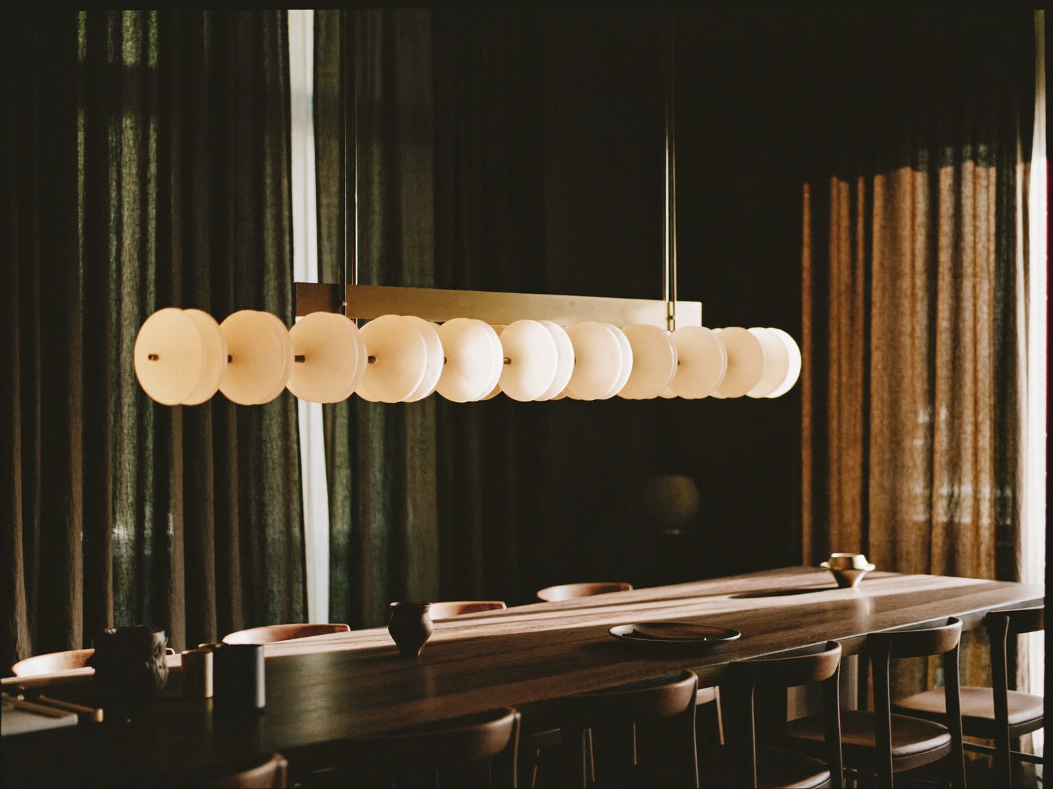 Sala da pranzo milanese con lampadario lineare in ottone e alabastro sopra un tavolo in noce scuro, luce calda di fine pomeriggio